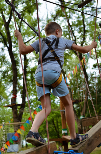  A child in equipment for climbers. A boy in an amusement park walks a tightrope with obstacles.