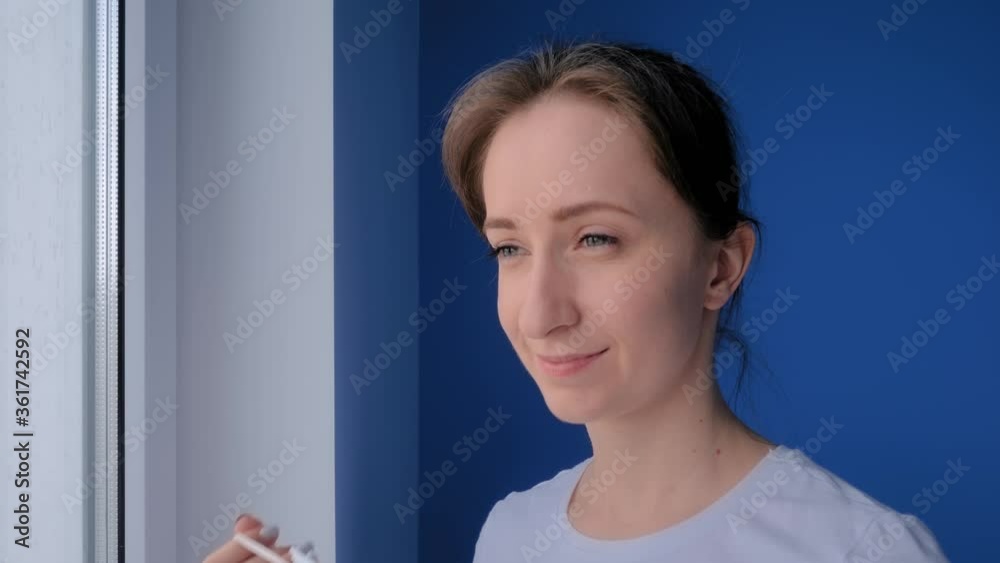 Slow motion: portrait of smiling , woman putting on medical face mask and looking out of window in room with blue wall. Self isolation, prevention, quarantine, COVID 19, coronavirus, safety concept Slow motion: portrait of smiling , woman putting on medical face mask and looking out of window in room with blue wall. Self isolation, prevention, quarantine, COVID 19, coronavirus, safety concept