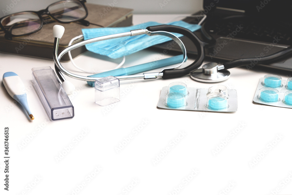 Workplace of a doctor. Laptop, stethoscope, pills, thermometer, protective mask in blue shades on a white table. Workplace of a doctor. Laptop, stethoscope, pills, thermometer, protective mask in blue shades on a white table.