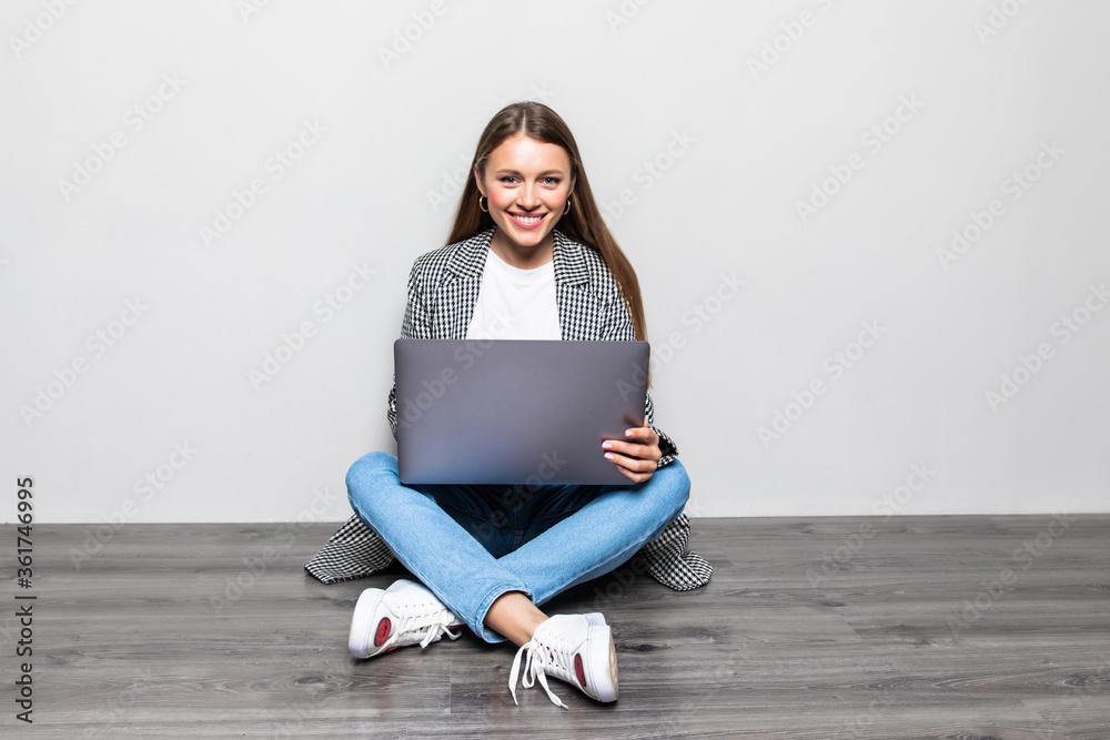 Happy young woman sitting on the floor with crossed legs and using laptop on gray background Happy young woman sitting on the floor with crossed legs and using laptop on gray background
