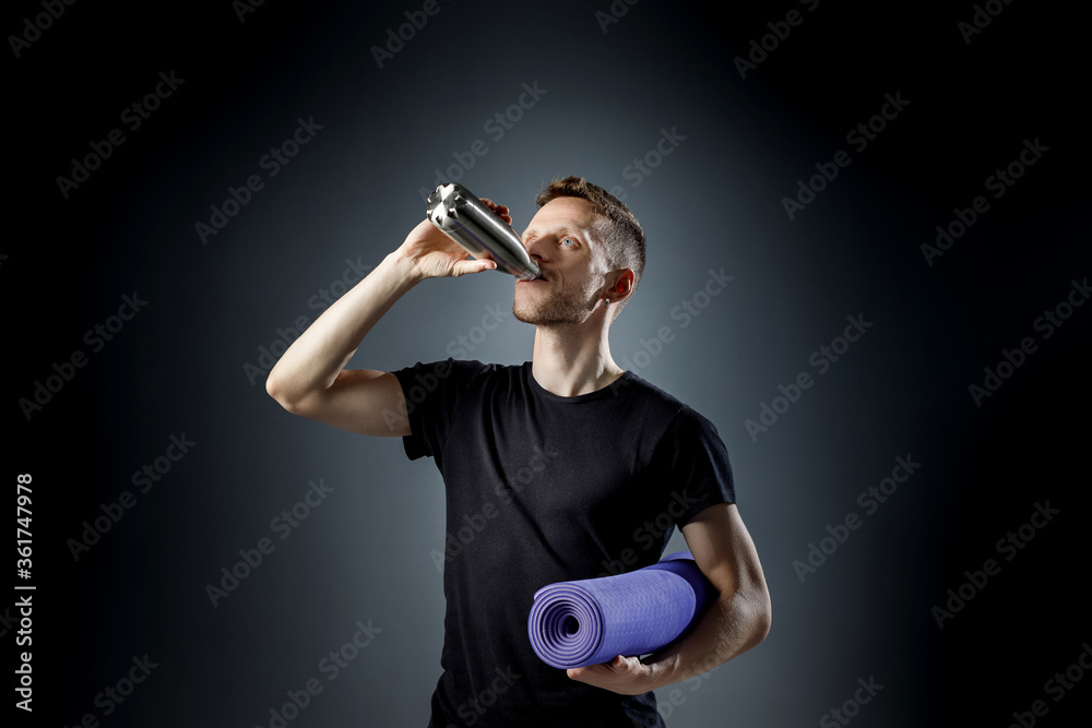 Young man on the black background is drinking water from fitness bottle, holding fitness mat Young man on the black background is drinking water from fitness bottle, holding fitness mat