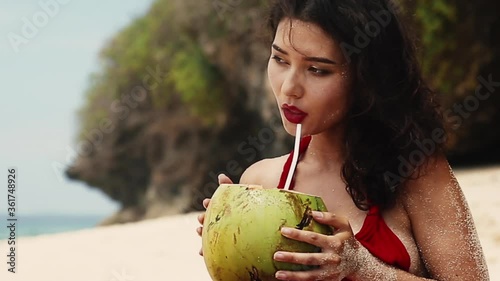 Beauty, baunty, travel, fashion concept.Beautiful pretty asian woman in hat and red dress drinking coconut on tropical paradise beach. Woman quench her thirst with coco nut. Vacation on exotic island photo