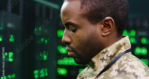 Close up of young African American army technician in camouflage unform standing at servers in secret monitoring military center and checking data. Militarian worker controlling digital facility. photo