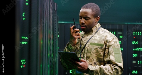 Obraz na plátně African American military man in uniform working in servers room, holding tablet device and speaking in walkie-talkie