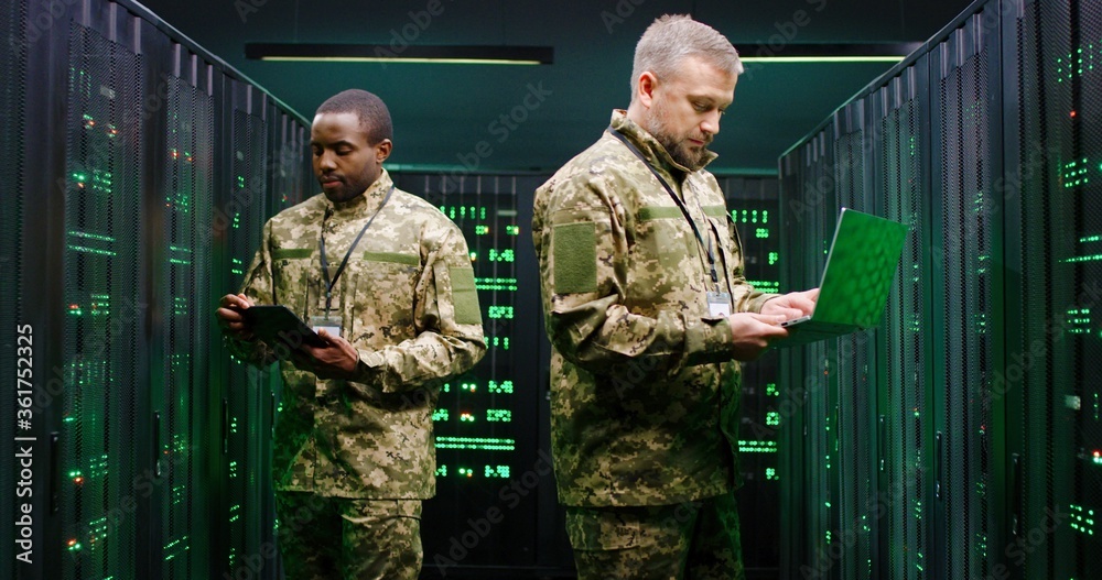 Fototapeta premium Multithnic military officials controlling servers and checking digital data in monitoring room. African American and Caucasian men colleagues with tablet device and lapptop computer. Wiretapping work.