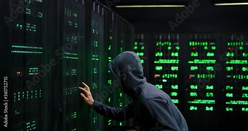 Male Asian guy data thief in dark hood stealing in the server room with tablet device in hands. Digital spy man hacking database. Male hacker commiting cyber crime.