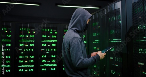 Asian male hacker hacking server system in dark sorage room with green lights. Police running in with pointing guns and arresting cyber criminal. FBI detention of governmental database thief.