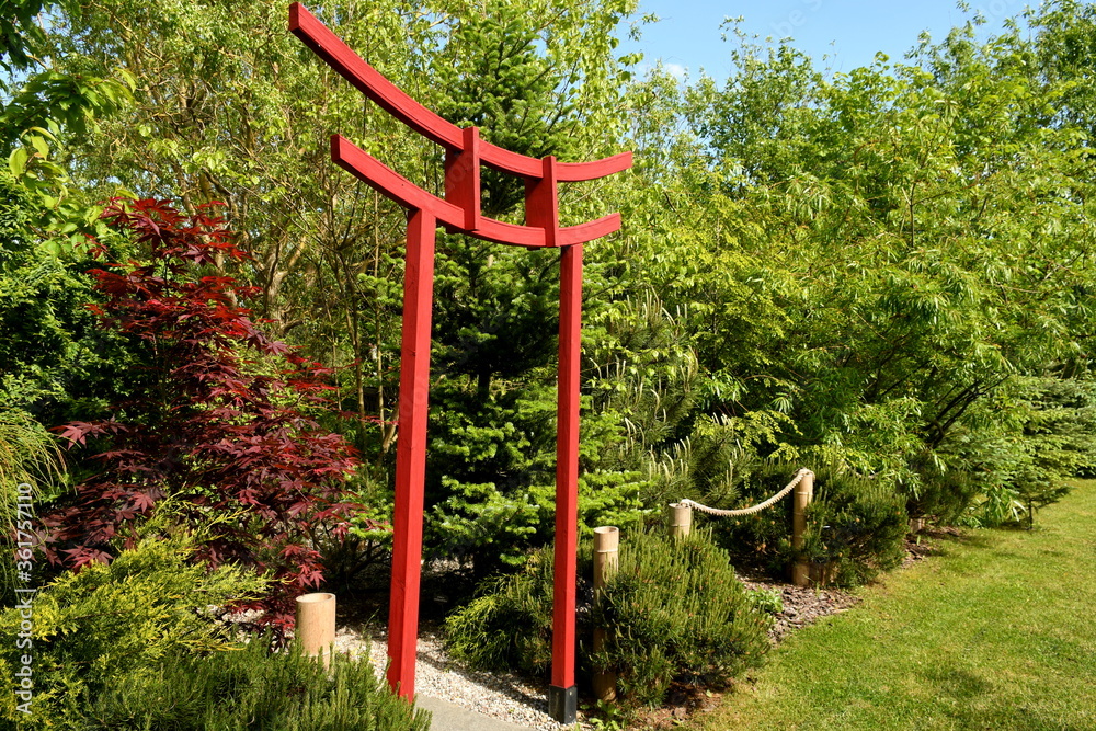 Close up on a red chinese style arch made out of wood and metal standing in the middle of a public park next to some shrubs, vegetation, and other flora seen on a sunny summer day in Poland