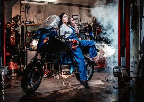 Hot brunette woman in blue overalls posing for a camera while leanign on sportbike in garage or workshop