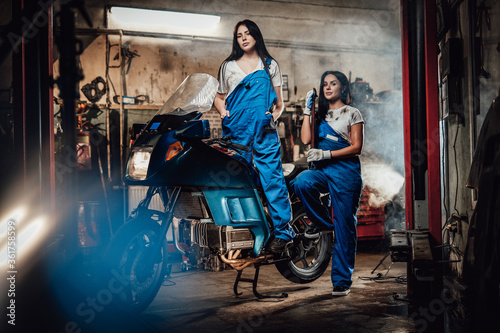 Two hot brunette women in blue overalls posing next to a sportbike in authentic workshop garage