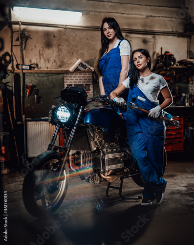 Two hot brunette women in blue overalls posing next to a custom bobber in authentic workshop garage