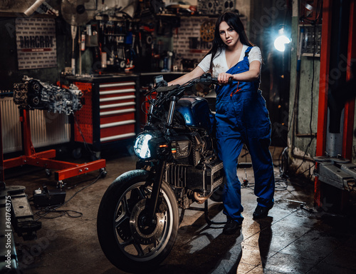 Young female mechanic in blue overalls posing with custom bobber in garage or workshop
