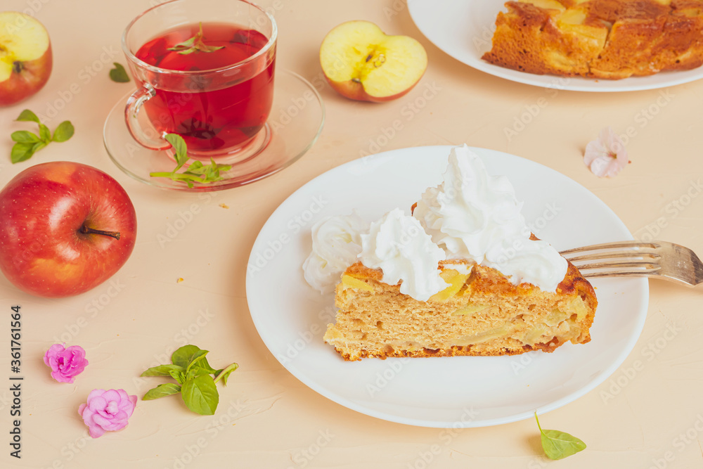 Charlotte apple cooked at home, garnished with whipped cream on a white plate. Around - apples, a cup of tea and tea leaves, flowers, mint leaves. A light image in pastel colors. Home baking concept. Charlotte apple cooked at home, garnished with whipped cream on a white plate. Around - apples, a cup of tea and tea leaves, flowers, mint leaves. A light image in pastel colors. Home baking concept.