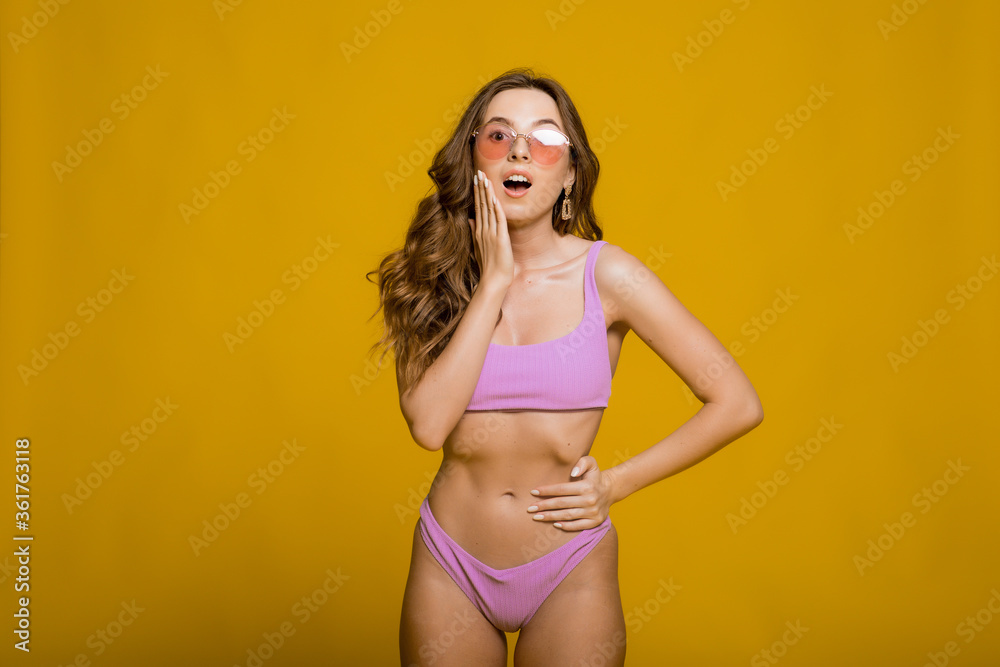 Beautiful young female in , sunglasses and swimsuit looking away while standing on yellow background. Copyspase - obrazy, fototapety, plakaty 