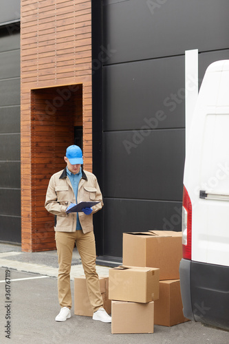 Young courier in uniform making notes in document he accepting parcels while standing outdoors near the warehouse