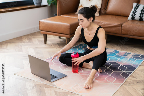beautiful Asian-looking girl sitting on a carpet in sportswear with a water bottle in her hands and a laptop at home 
