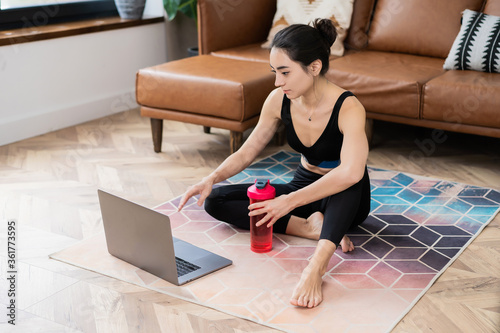 beautiful Asian-looking girl sitting on a carpet in sportswear with a water bottle in her hands and a laptop at home
