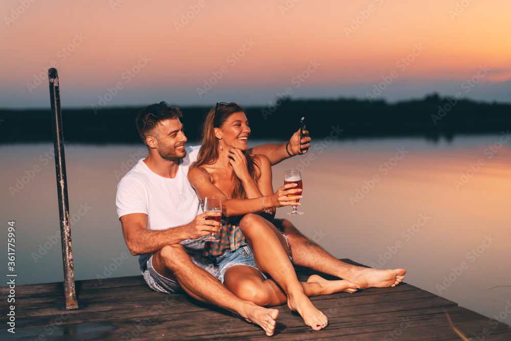 Young couple sitting on the dock and taking selfies during summer evening