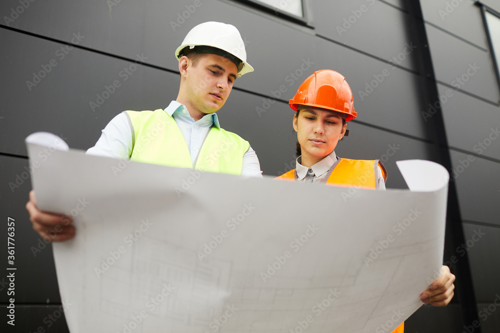 Man and woman in work helmets holding blueprint of new building and examining it in team while standing outdoors
