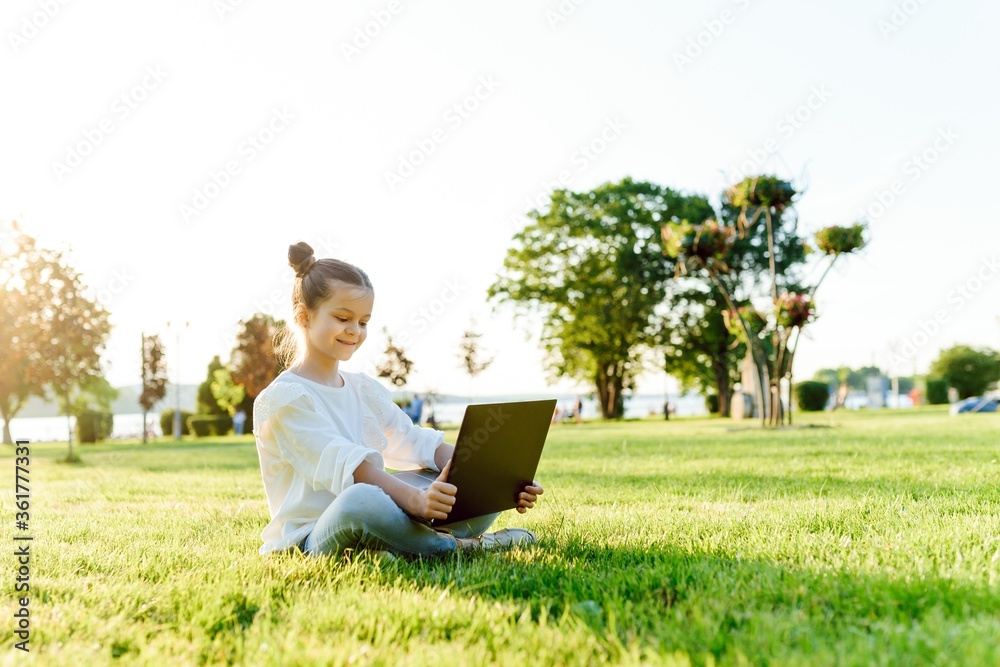 © Serhii - Little girl sitting in the park and working with laptop. Education, lifestyle, technology concept