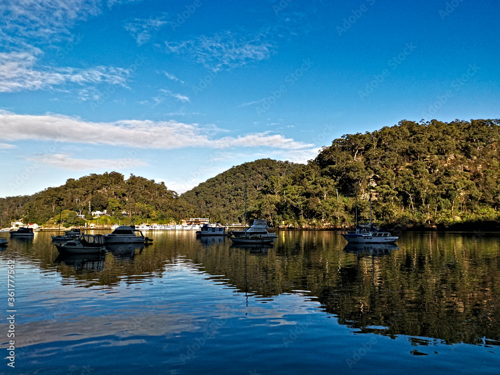 Beautiful morning view of Creek with boats and reflections of blue sky, light clouds, mountains and trees. Berowra Waters, Berowra Valley National Park, New South Wales, Australia
