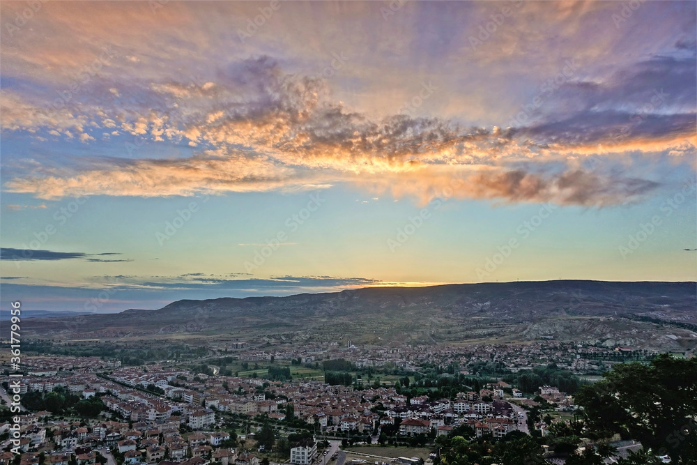 Dawn in the valley of Cappadocia. The sun rises from behind the mountains on the horizon. The sky is blue, the clouds are painted in pink, gold, lilac tones. In the valley is a village .