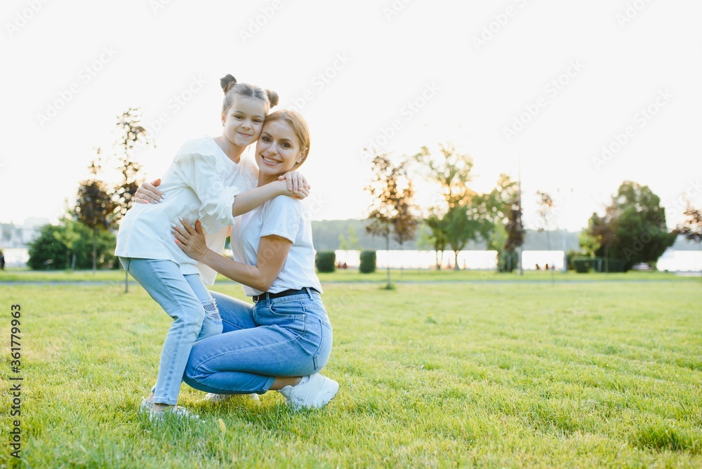 Fototapeta premium Attractive young woman with her little cute daughter are spending time together outdoors. Mom with daughter in park on a green grass during the sunset.
