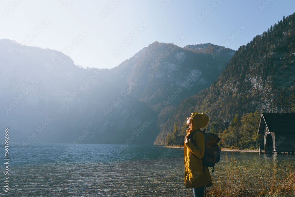 side view of a girl in a yellow autumn jacket with a backpack in the mountains looks at the sun on the background of the lake