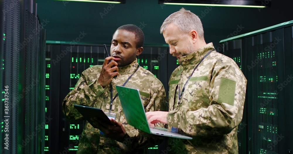 Fototapeta premium Multiethnic male army officers with tablet device and laptop computer checking secret data in server room and passing via walkie-talkie. African American and Caucasian men talking at digital facility.