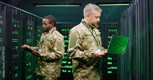 Mixed-races male military co-workers operating with data in server center and checking information with tablet device amd laptop computer. African American and Caucasian men working with servers.
