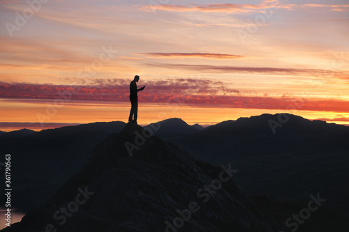 A silhouette of man using smart phone on the top of mountain against the backdrop of a stunning sunset sky. Loch Lomond and The Trossachs National Park, Scotland