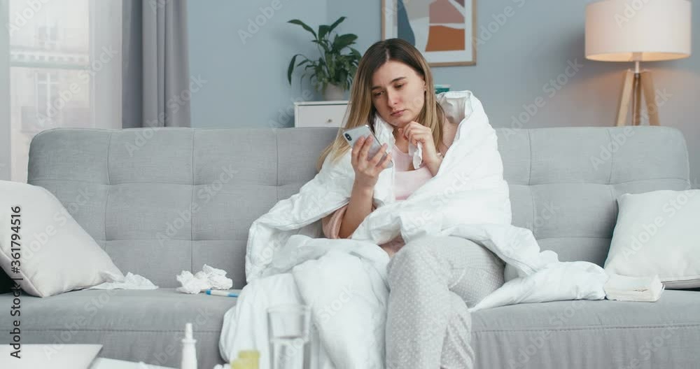 Caucasian young ill female using smartphone while sitting on couch in blanket, caughing and blowing nose in napkin. At home. Unhealthy girl tapping and scrolling on mobile phone on sofa. Caucasian young ill female using smartphone while sitting on couch in blanket, caughing and blowing nose in napkin. At home. Unhealthy girl tapping and scrolling on mobile phone on sofa.