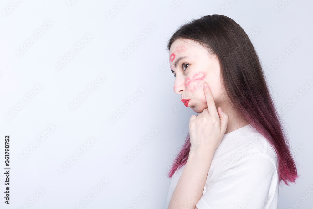 Happy young girl with kisses imprints on her face with red lipstick. Shows finger place to kiss on the cheek. World Kissing Day Concept. Copy space Happy young girl with kisses imprints on her face with red lipstick. Shows finger place to kiss on the cheek. World Kissing Day Concept. Copy space