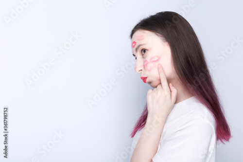 Happy young girl with kisses imprints on her face with red lipstick. Shows finger place to kiss on the cheek. World Kissing Day Concept. Copy space