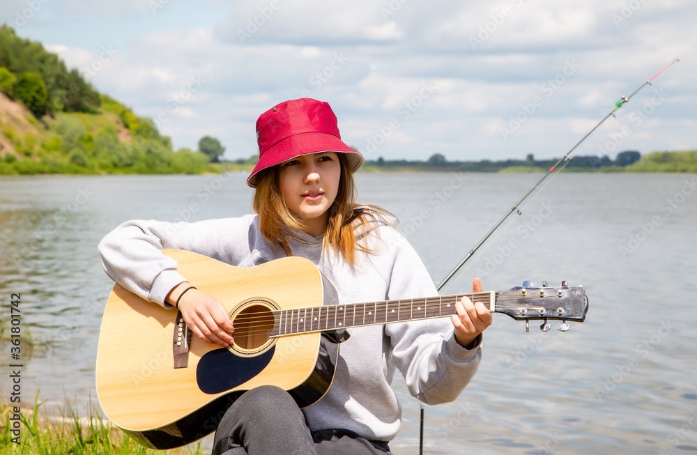 young beautiful girl in red panama plays guitar on the river