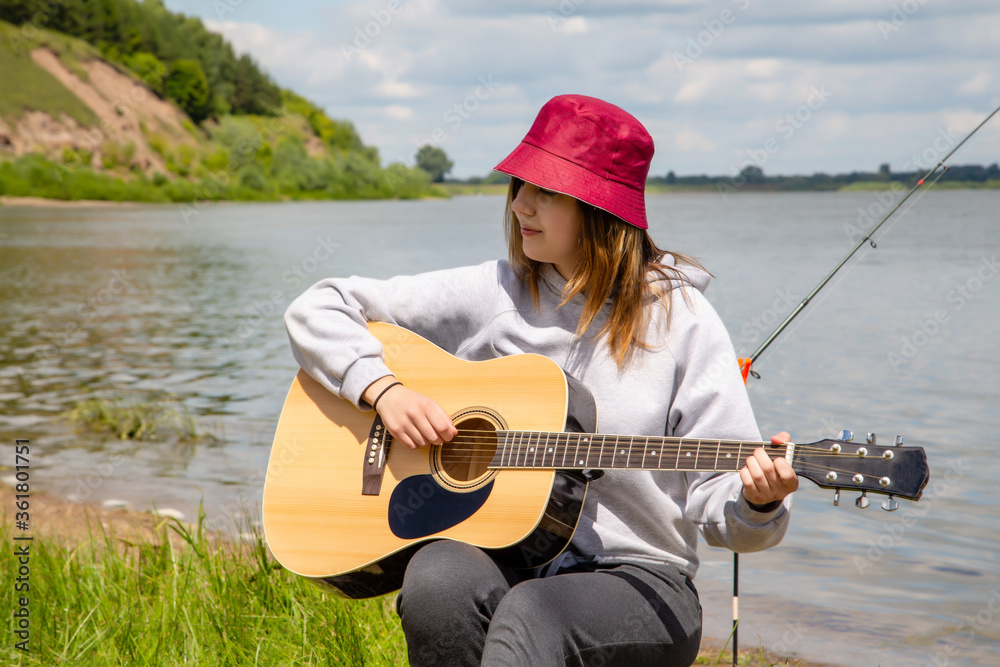 young beautiful girl in red panama plays guitar on the river