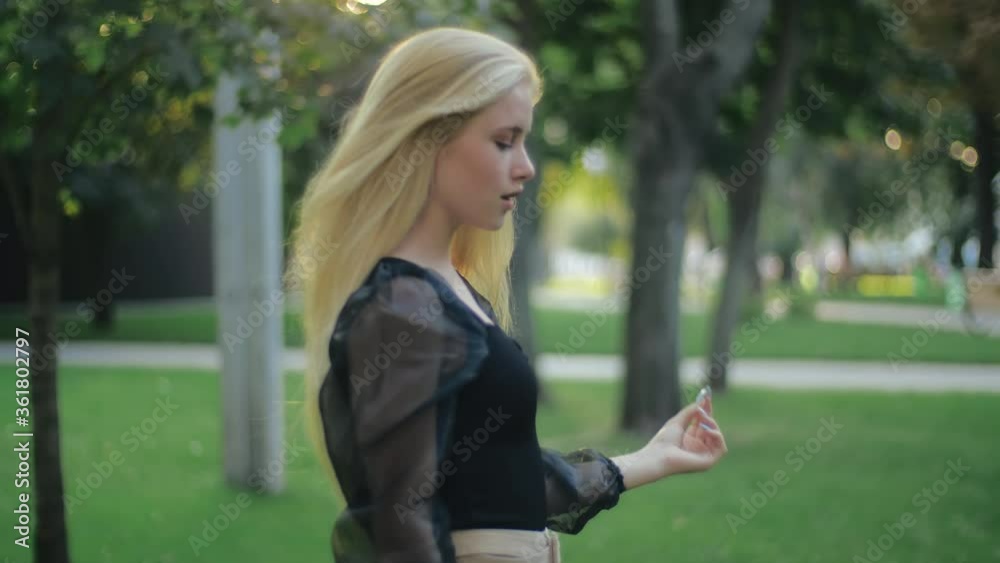 A young attractive girl smiles happily and walks in the park, spins beautifully, straightens her long blond fluffy hair, highlighted by the sun. A model looks at the camera and poses in green leaves. A young attractive girl smiles happily and walks in the park, spins beautifully, straightens her long blond fluffy hair, highlighted by the sun. A model looks at the camera and poses in green leaves.