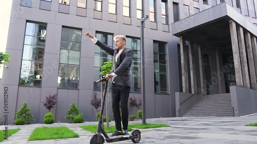 Attractive satisfied stylish young light-haired guy enjoying his ride on own e-scooter near beautiful architectural building photo