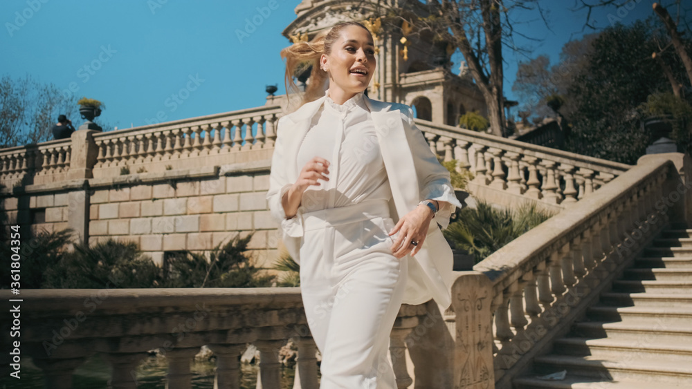 Young attractive elegant woman in classic white suit happily running down the stairs in old park