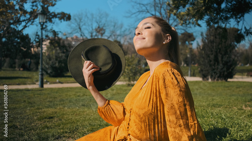 Young beautiful woman in bright orange overalls with hat dreamily resting on lawn in city park. Pretty model posing outdoor under the sun photo