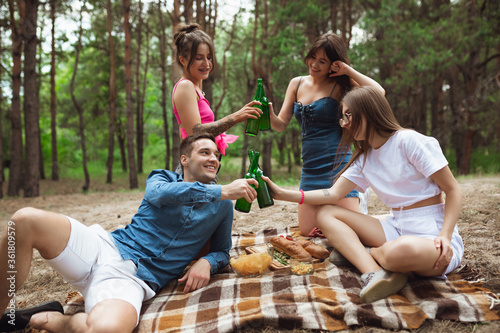 Happiness. Group of friends clinking beer bottles during picnic in summer forest. Lifestyle, friendship, having fun, weekend and resting concept. Looks cheerful, happy, celebrating, festive.