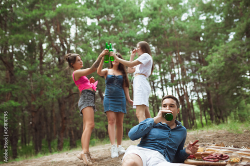 Togetherness. Group of friends clinking beer bottles during picnic in summer forest. Lifestyle, friendship, having fun, weekend and resting concept. Looks cheerful, happy, celebrating, festive.