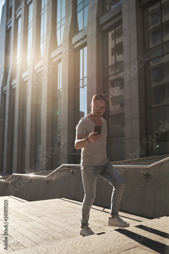 a stylish guy, wearing glasses, climbs the steps to the office check-in with a phone in his hands. light jeans and a fashionable gray T-shirt. beautiful contour sunlight