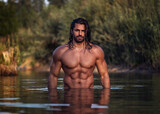Long haired bearded muscular man shirtless stands waist deep in the water. Aquaman in the water. Tanned attractive guy with long dark wet hair