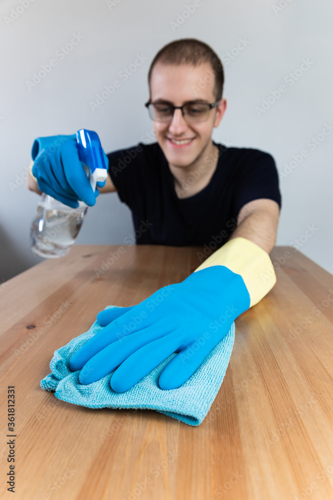 Young man cleaning a wooden table with blue cloth and alcohol sanitizer, wearing blue gloves, glasses and smiling. Vertical Young man cleaning a wooden table with blue cloth and alcohol sanitizer, wearing blue gloves, glasses and smiling. Vertical
