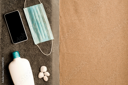 covid-19 prevention beach concept, cris towel on the beach sand, smartphone with black screen, blue virus protection mask, cream bottle and white stones. Sandy beach. Top view. space for text