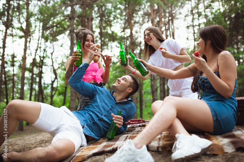 Youth. Group of friends clinking beer bottles during picnic in summer forest. Lifestyle, friendship, having fun, weekend and resting concept. Looks cheerful, happy, celebrating, festive.