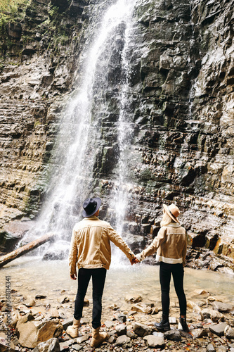 Young and beautiful couple at the mountain waterfall - Happy tourists visiting mountains. Lovestory. Tourists in hats. Military fashion.