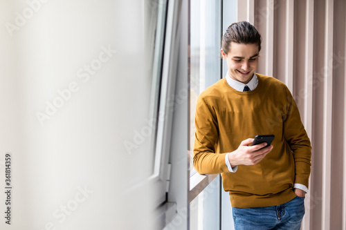 Enjoying office life. Joyful young man holding mobile phone and looking at camera on office background