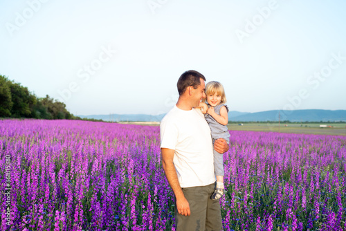 A smiling father holds his daughter in his arms, they laugh cheerfully on a walk in the purple field. Harmonious relationship between father and daughter.